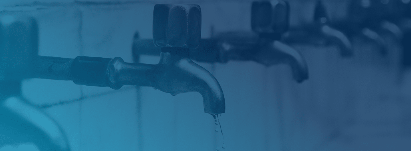 A row of black water taps mounted on a tiled wall, with one tap in the foreground slightly dripping water. The image has a blue overlay.