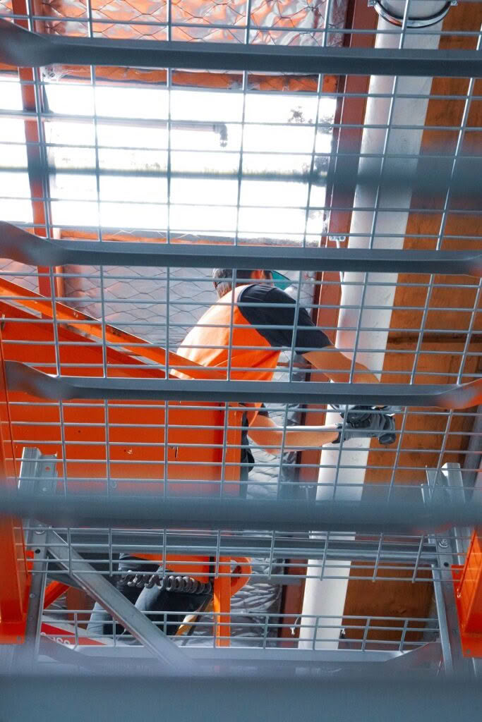 View from below of a person in an orange vest climbing a metal staircase, with metal grating and beams visible above and around them. The angle emphasizes the industrial structure.